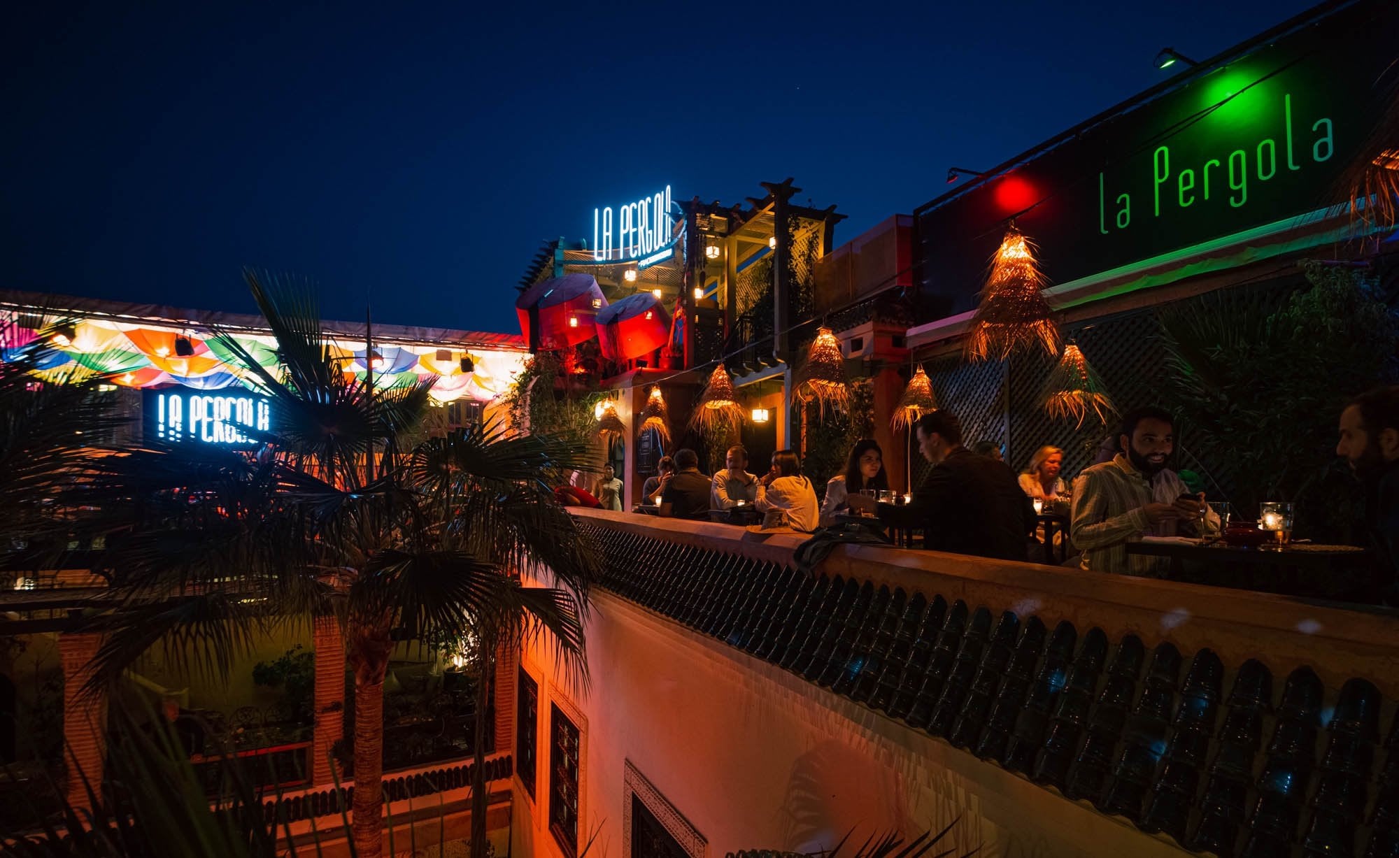 La Pergola rooftop at Riad Monceau at dusk, guests at tables under neon signs and palm trees