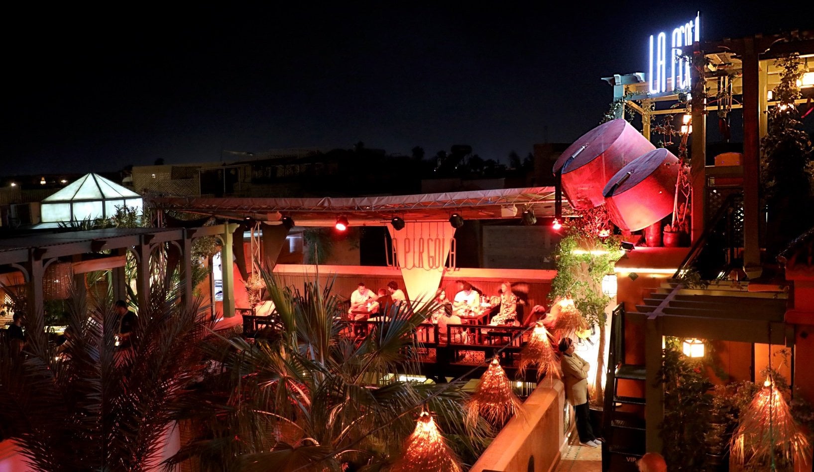 La Pergola rooftop at night, diners on a lit terrace among palms and neon signs