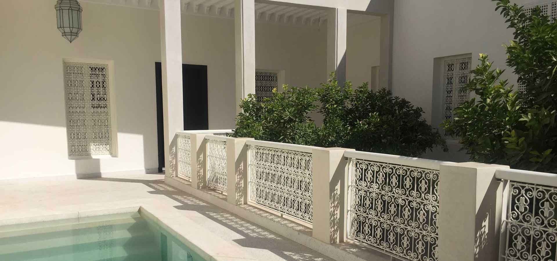 Sunlit plunge pool at Numa Marrakech beside a white gallery with wrought-iron railings and orange trees