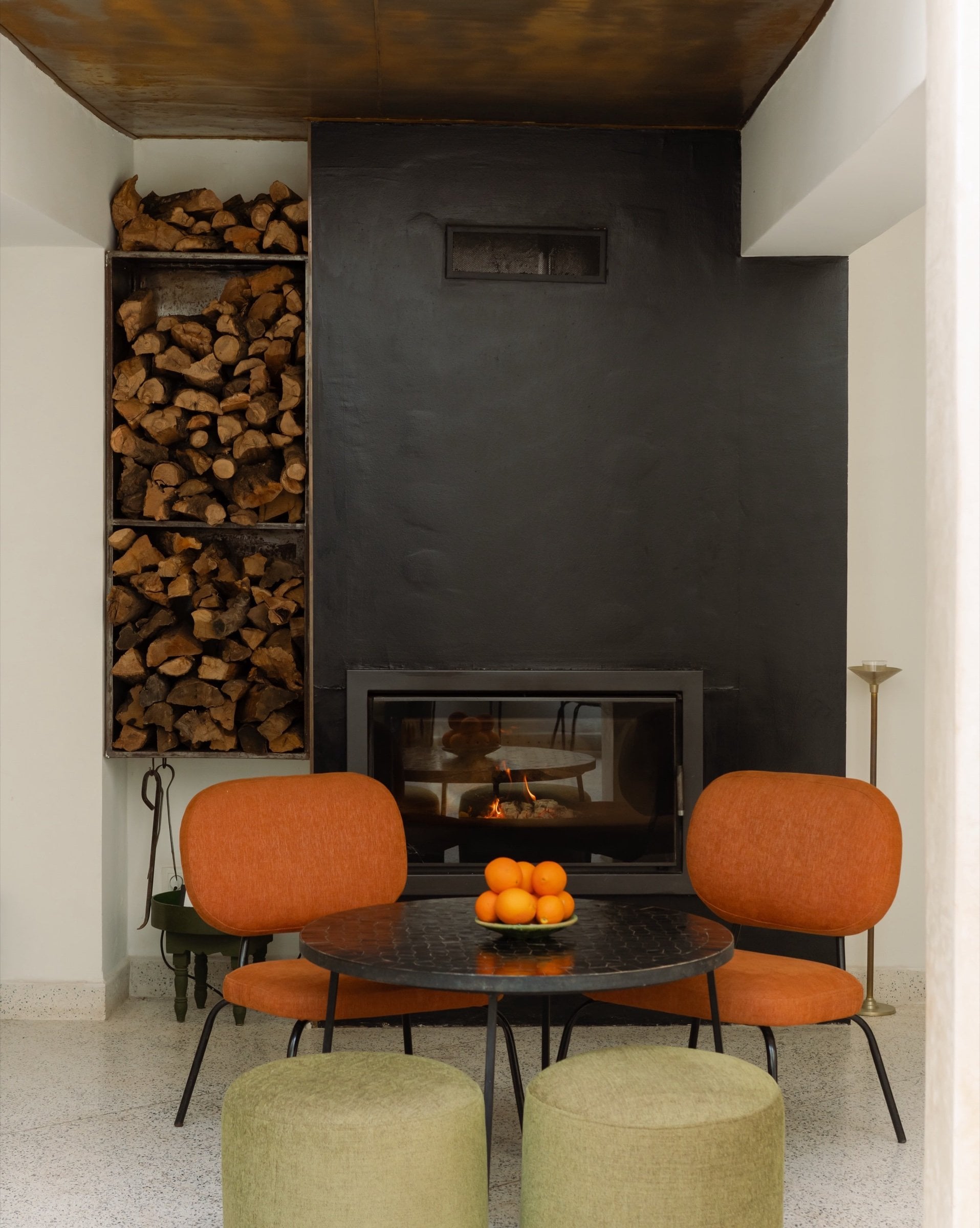 Fireplace corner in the patio of Numa Marrakech, with orange armchairs, a bowl of oranges and firewood