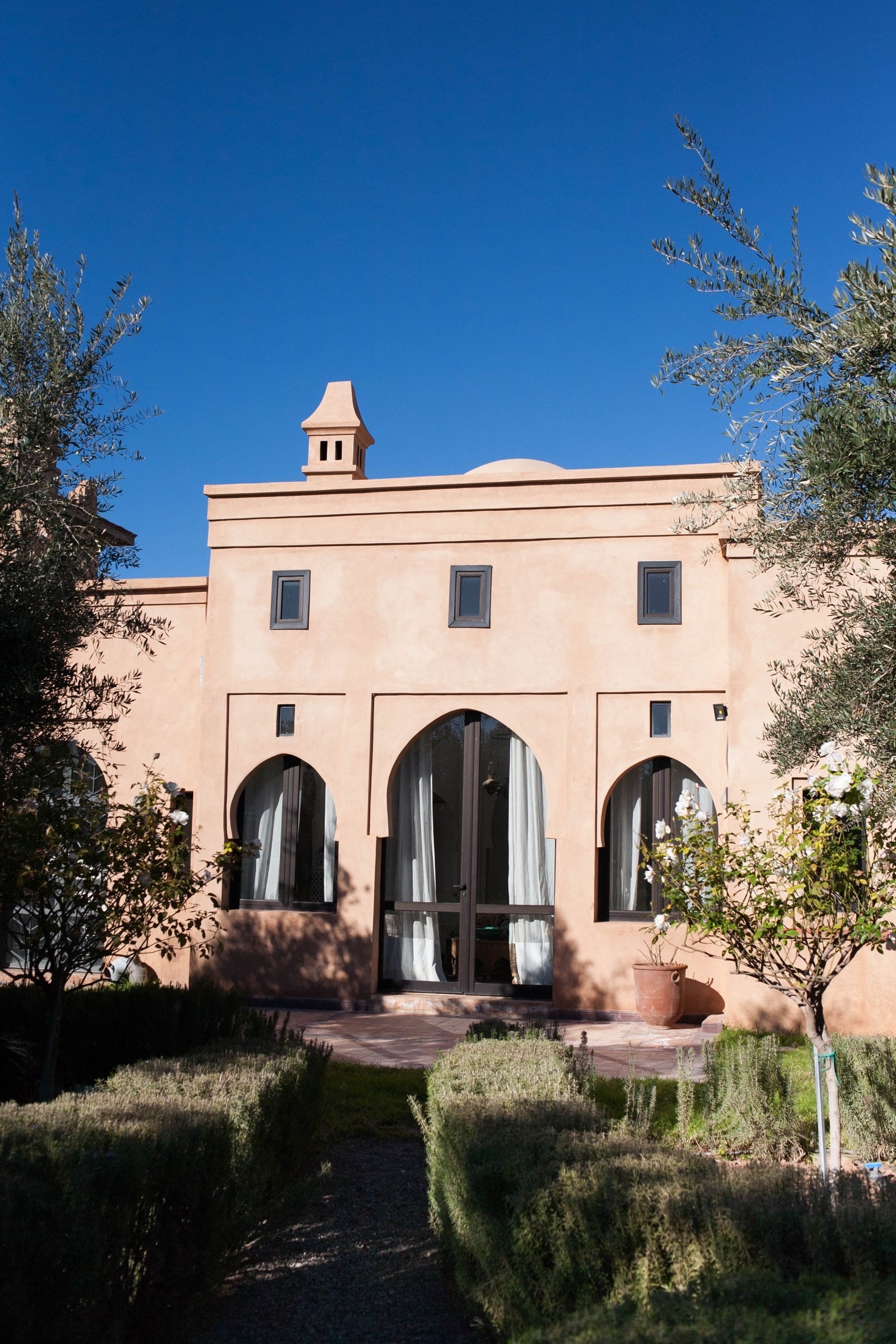 Medina Pavilion facade along an olive-lined stone path, terracotta walls and a stepped chimney against a blue sky.