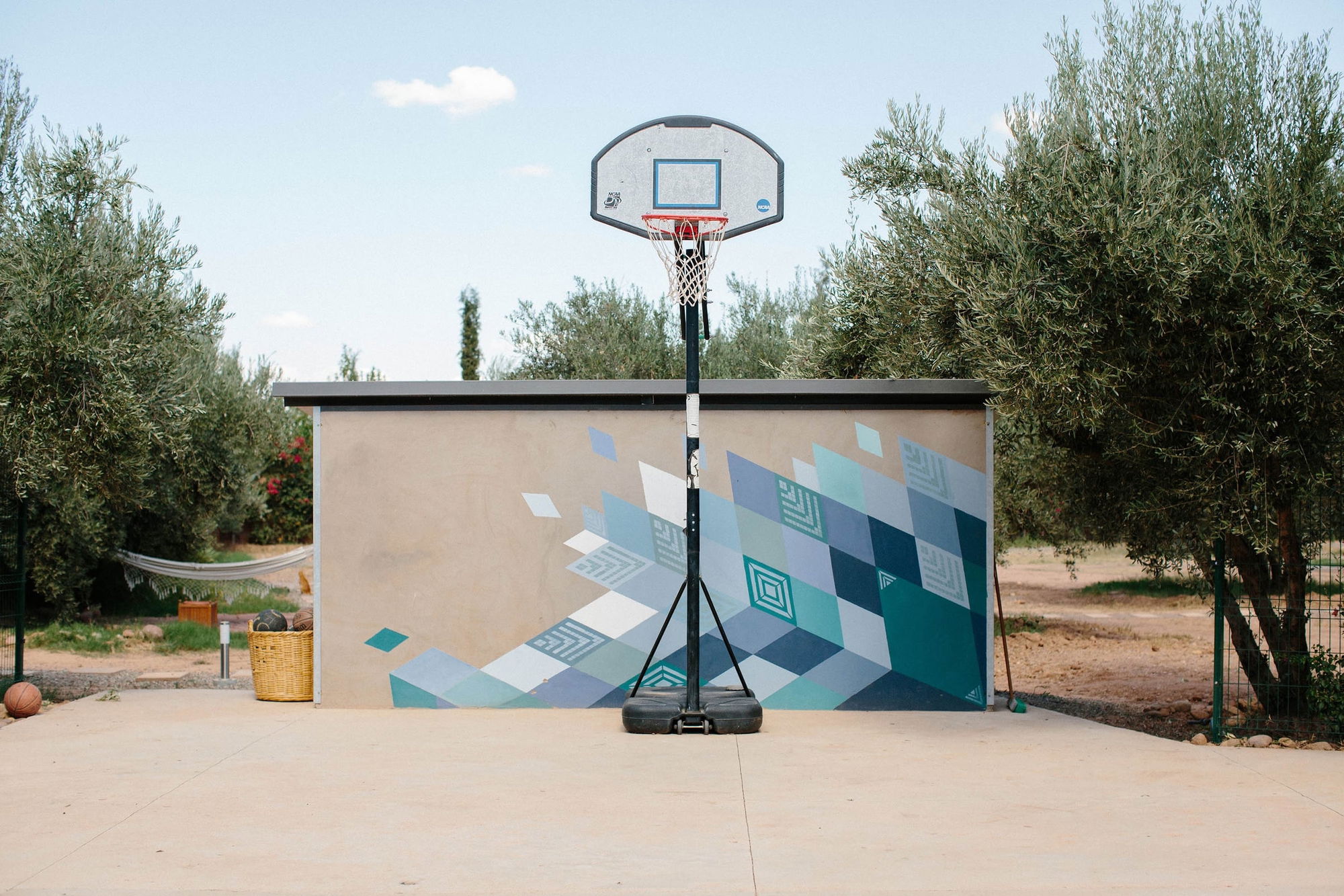 Outdoor basketball court with a geometric teal and blue mural on the backboard wall, set inside the olive grove.