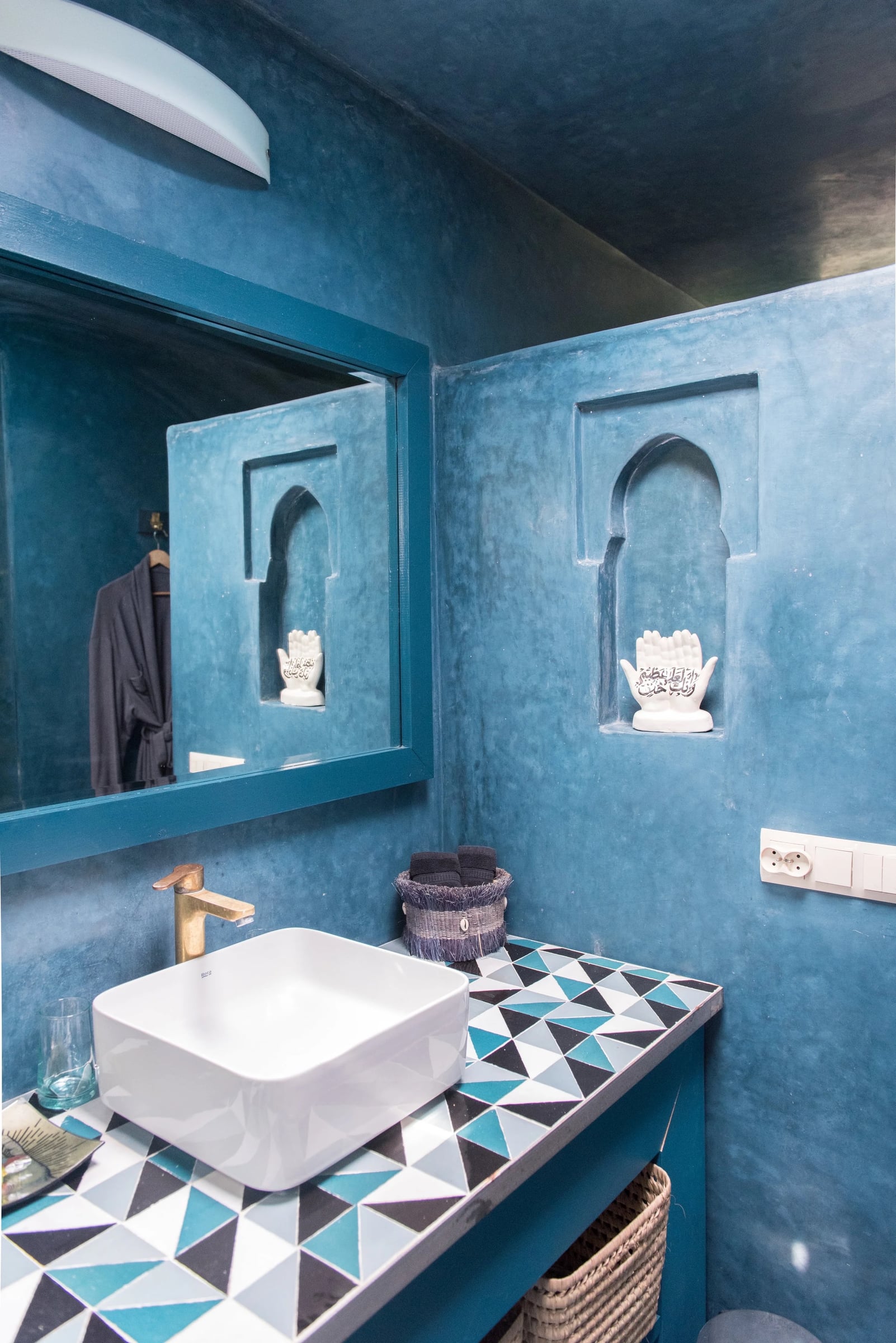 Bungalow washroom in burnished blue plaster with a triangle-mosaic vanity top and a carved arched wall niche.