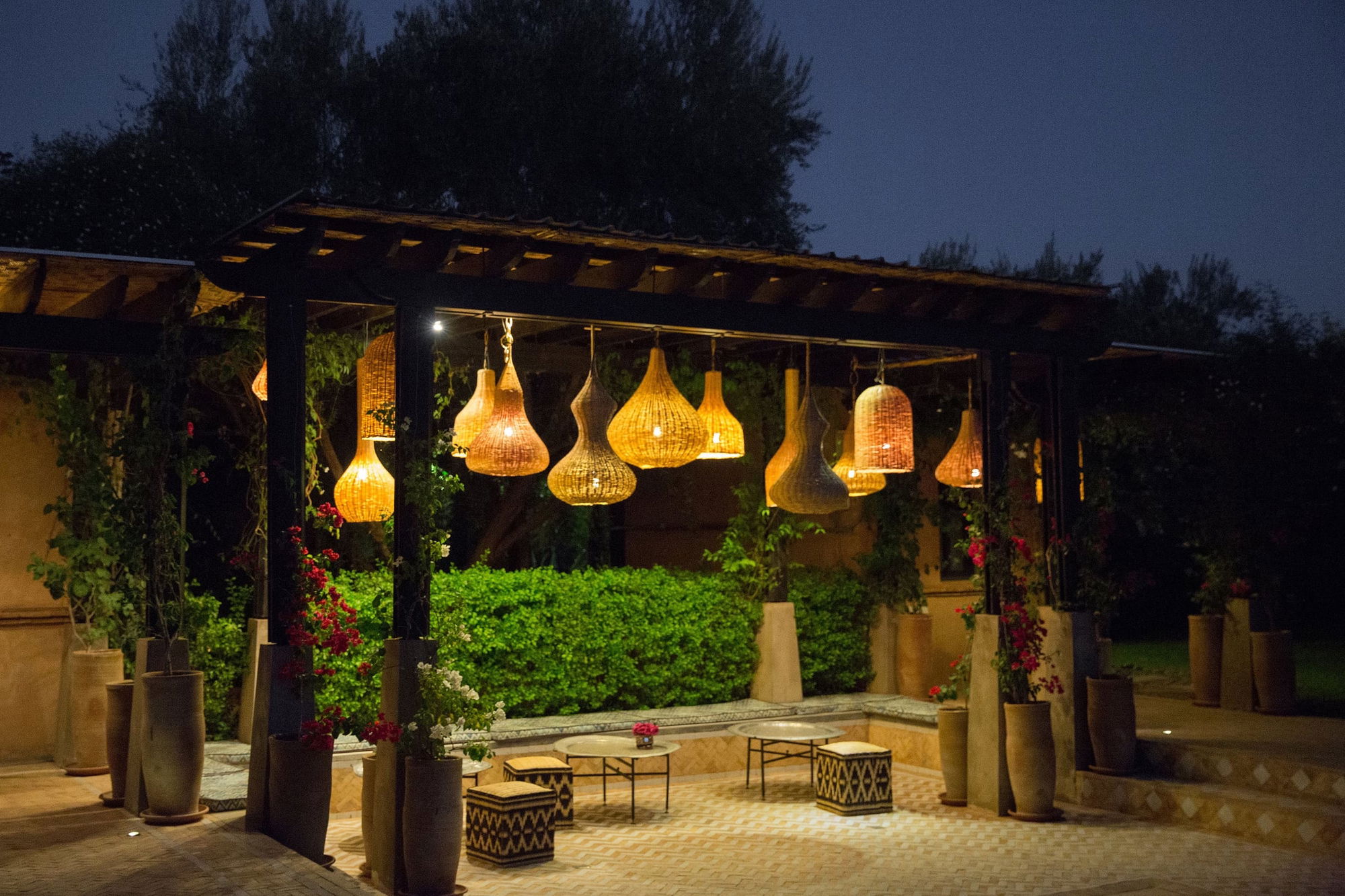 Poolside pergola lit at night with a cluster of woven pendant lamps over bougainvillea and stone benches.