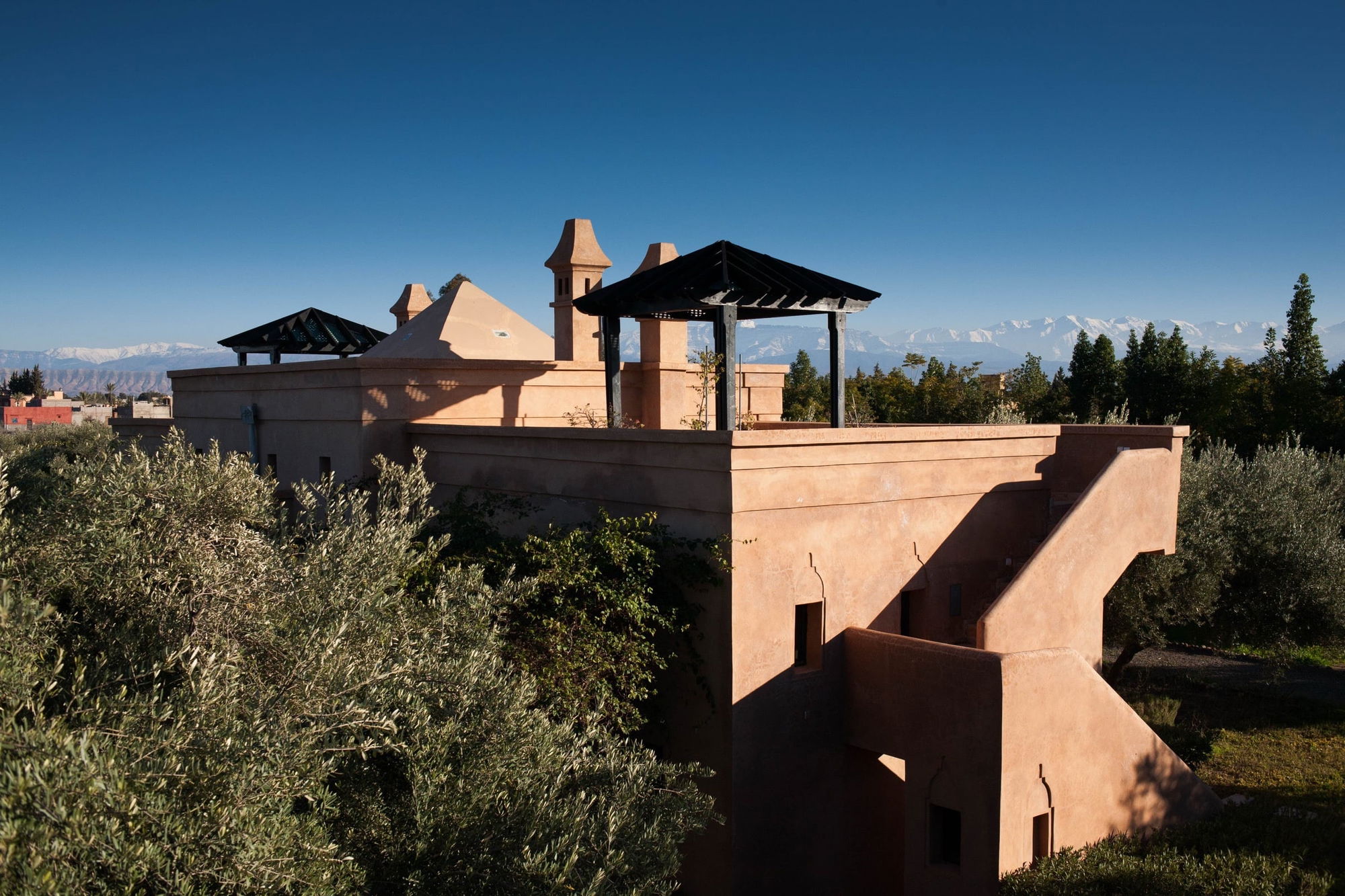 Rooftop of the Atlas Pavilion with a pyramid-roofed pavilion and chimneys, olive trees and snow-capped Atlas Mountains beyond.