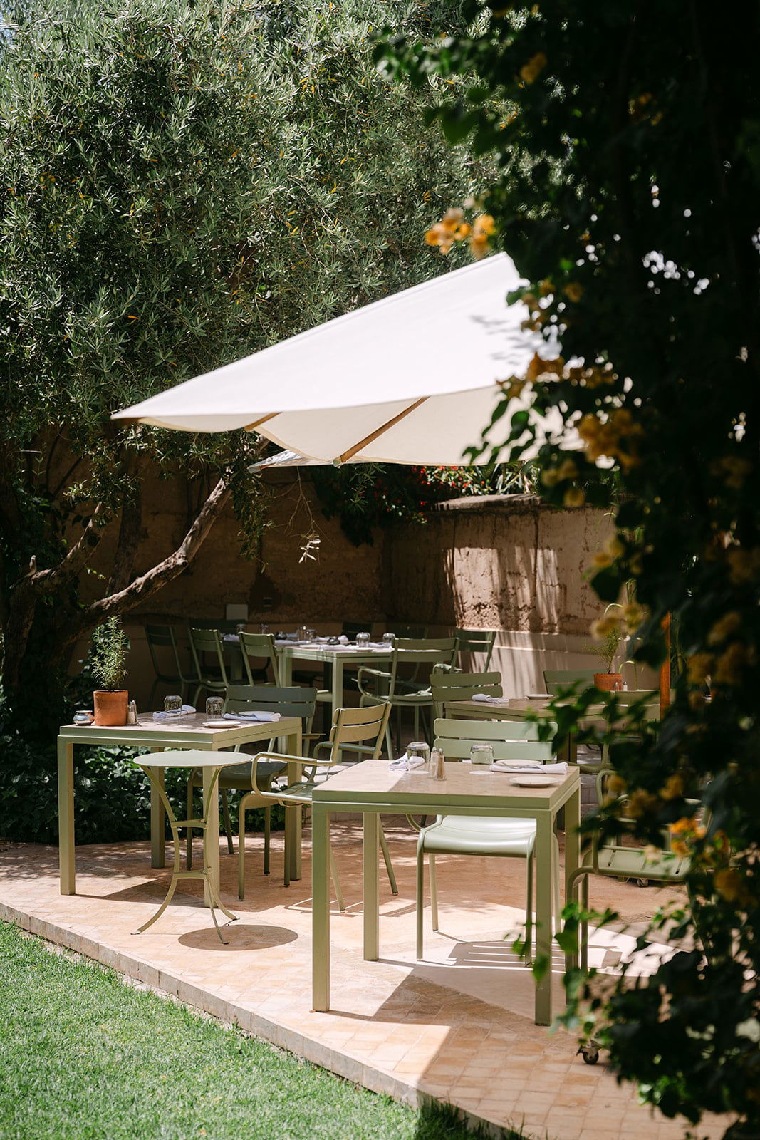 Open-air garden terrace laid for lunch, sage-green tables and chairs shaded by olive trees and a white parasol.