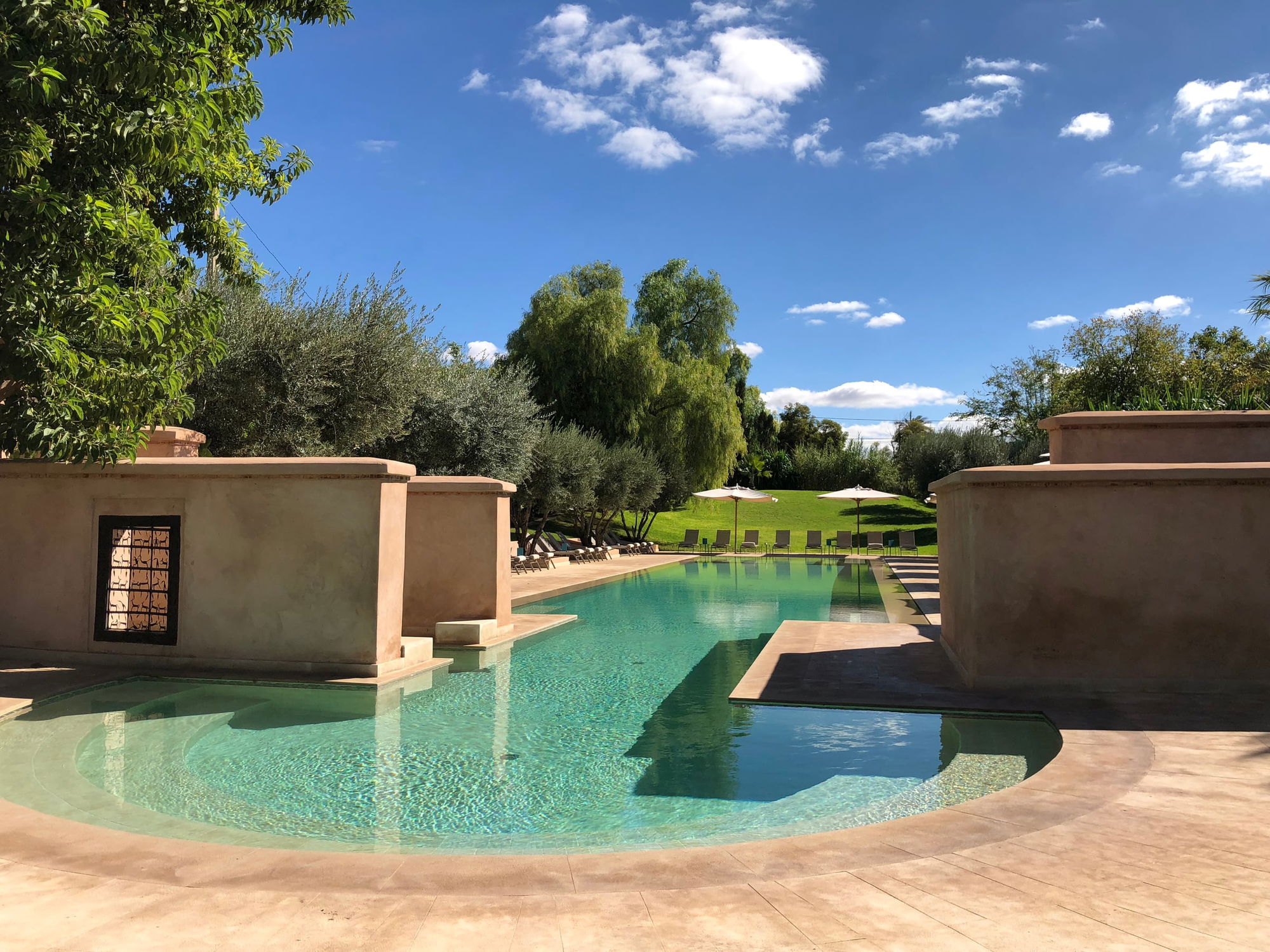 Curved main pool at Les Deux Tours, ochre walls on one side, loungers and white parasols on the lawn beyond.