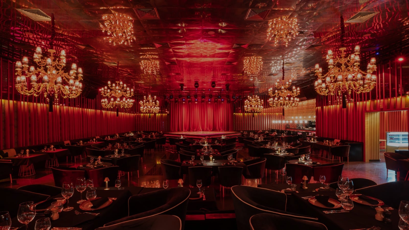 Main dining room in wide shot: red fluted wall panels floor to ceiling, mirrored ceiling with tiered gold chandeliers, round stage with a red velvet curtain, black tables set in the foreground.