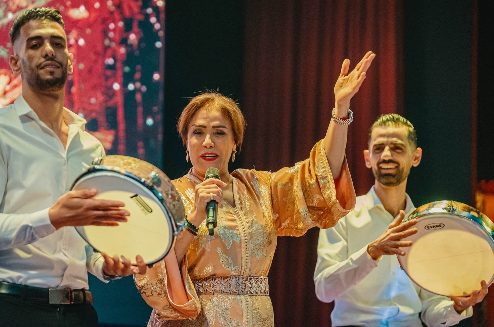 Singer in a gold caftan with a microphone between two musicians in white shirts holding frame drums, red curtain behind.