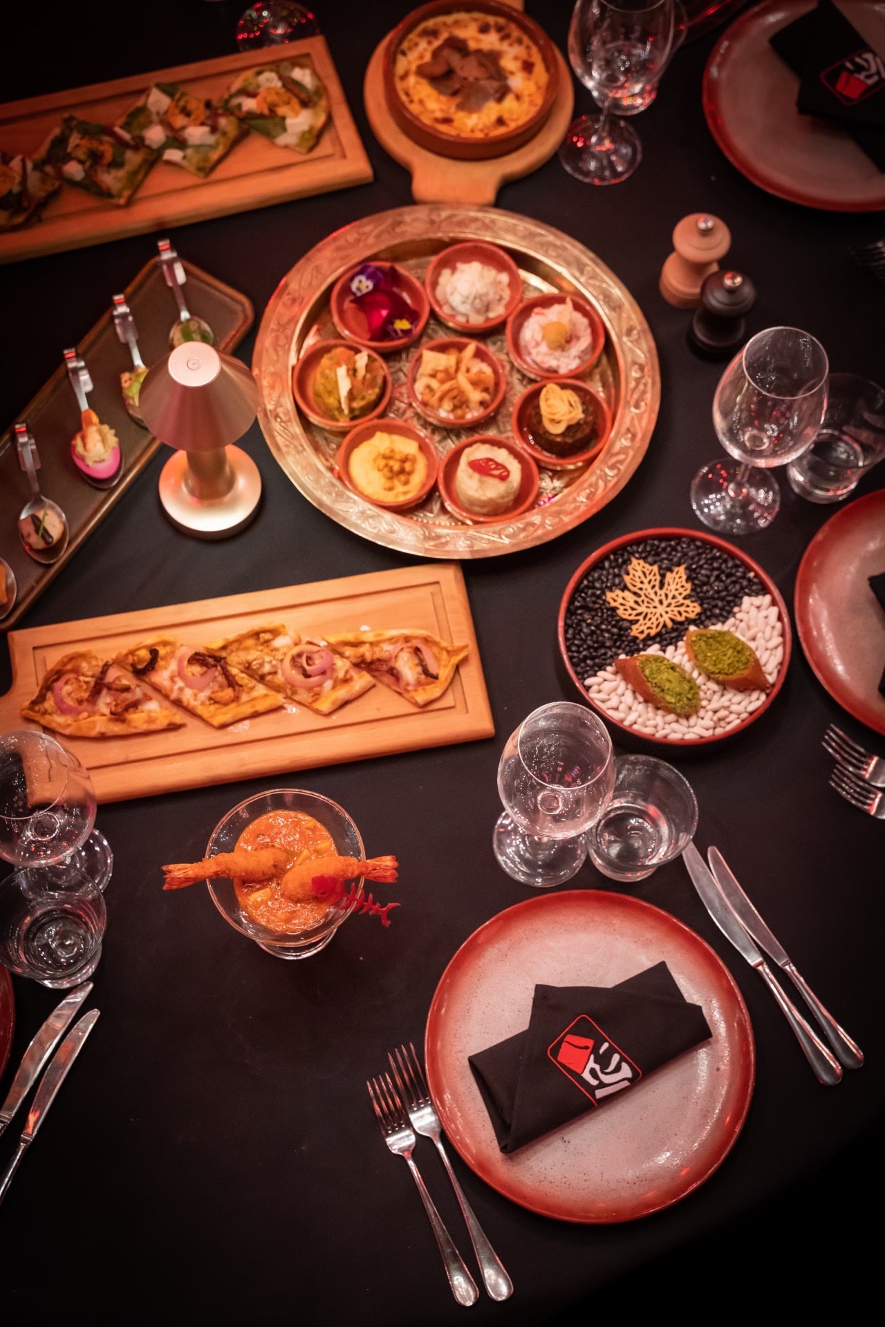 Table from above: round copper tray with nine small dishes, flatbread boards, prawn cocktail, black napkin with the venue logo.