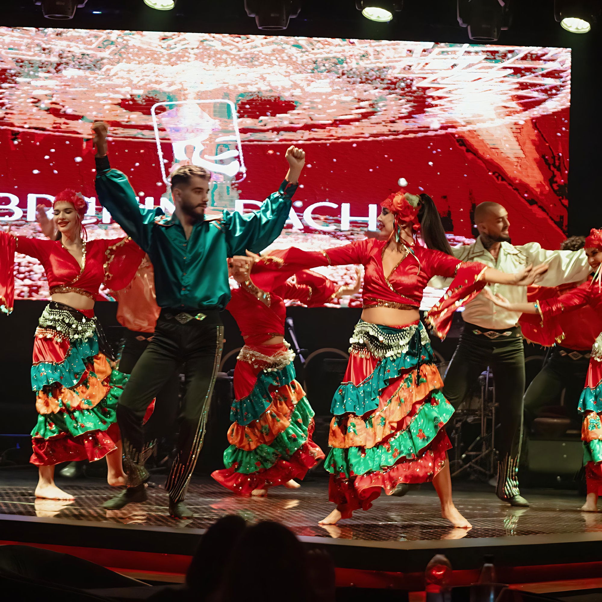 Dance company in red, green and gold costumes on stage, IBRAHIM PACHA on the LED screen behind.