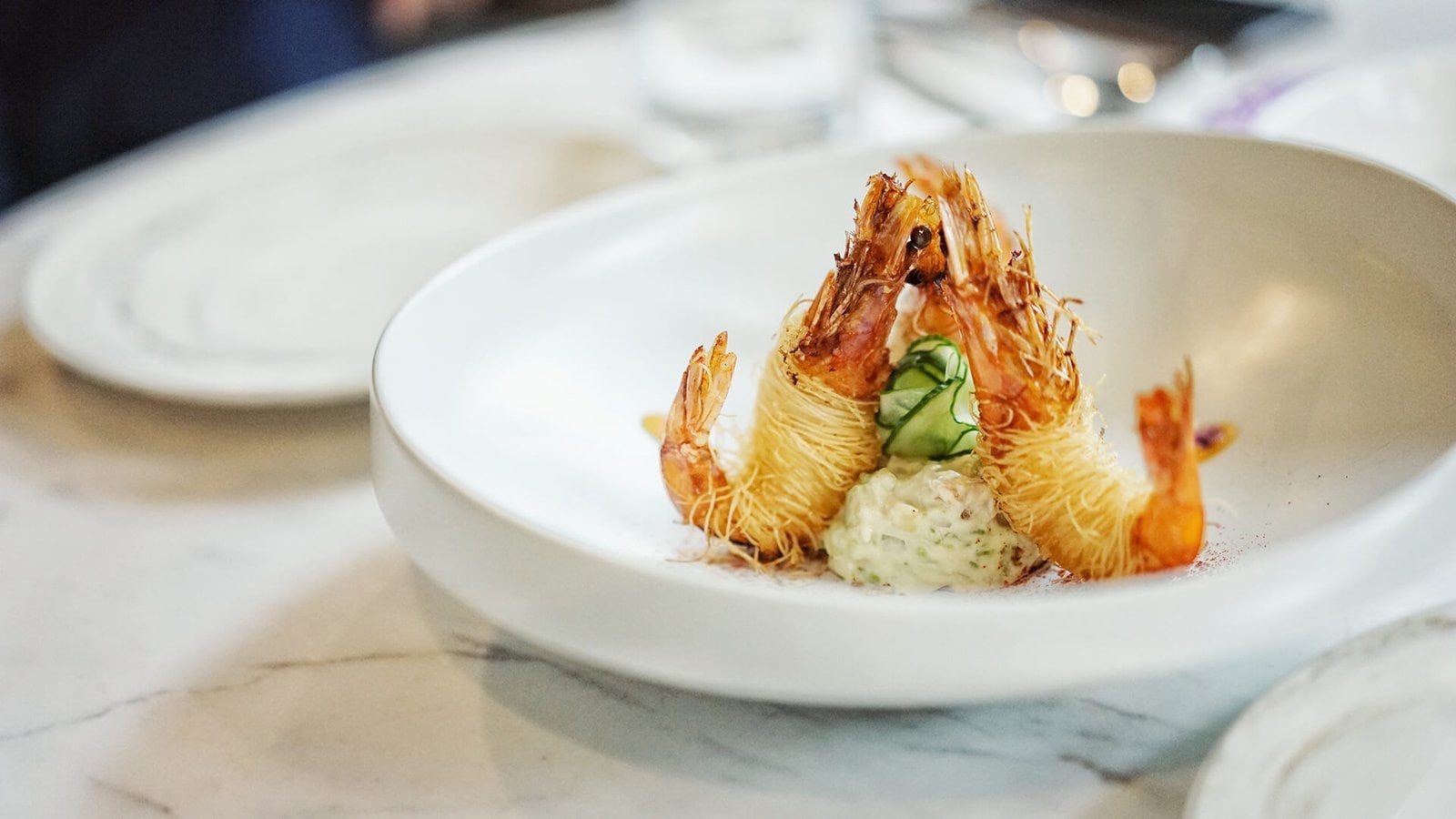 Three prawns wrapped in crisp kataifi on a herb and cucumber cream, marble table.