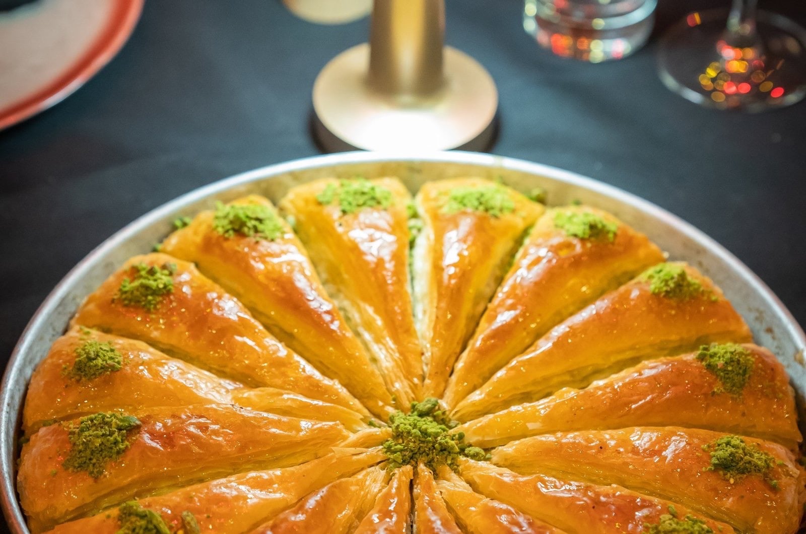 Round tray of baklava with pistachio, table lamp and a glass behind.