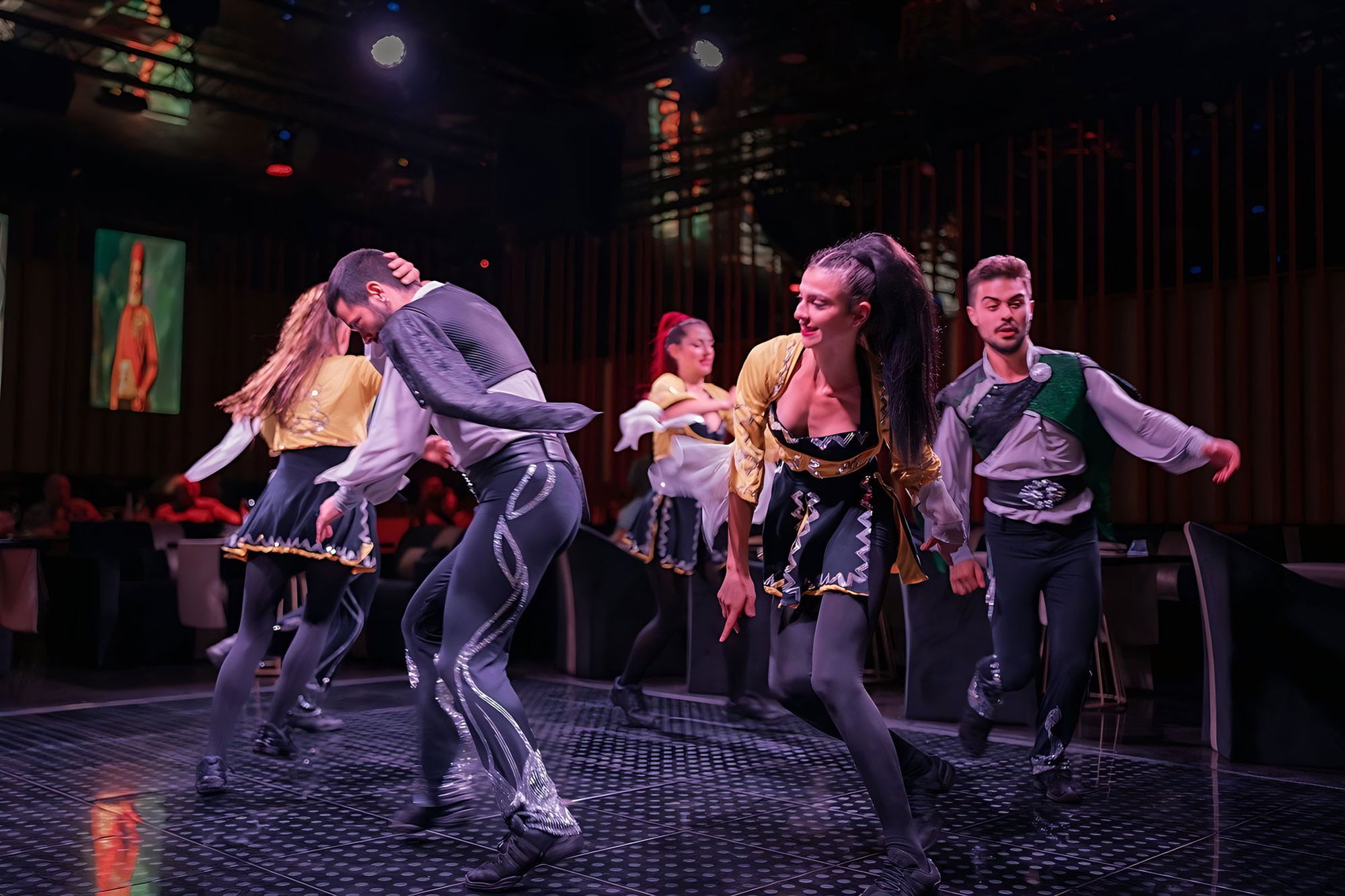 Dance company in dark and gold costumes moving between the tables, red fluted panels behind.