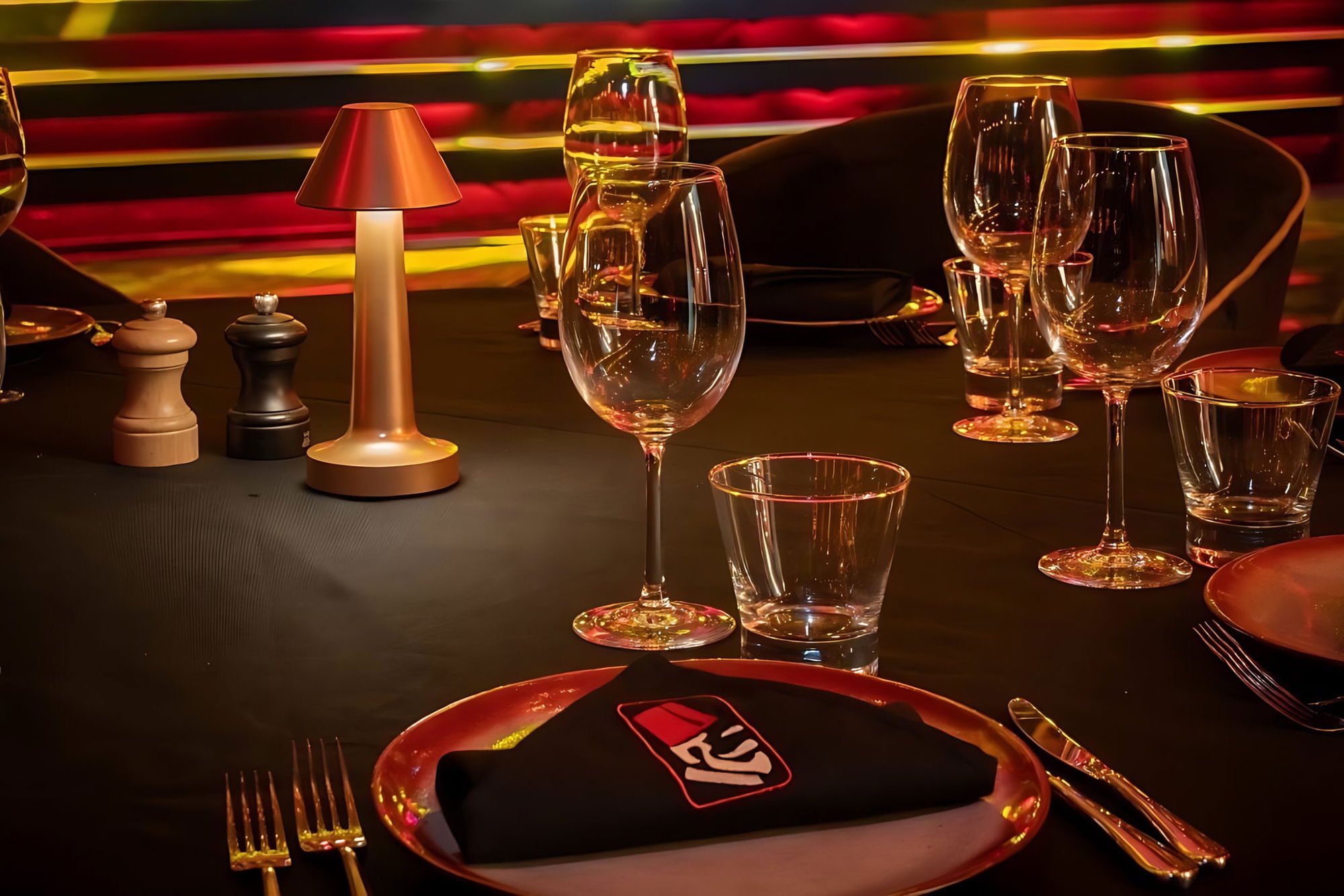 Table setting in close-up: a gold cordless table lamp, wine and water glasses, salt and pepper mills, black napkin with the red and black diamond logo of the venue.