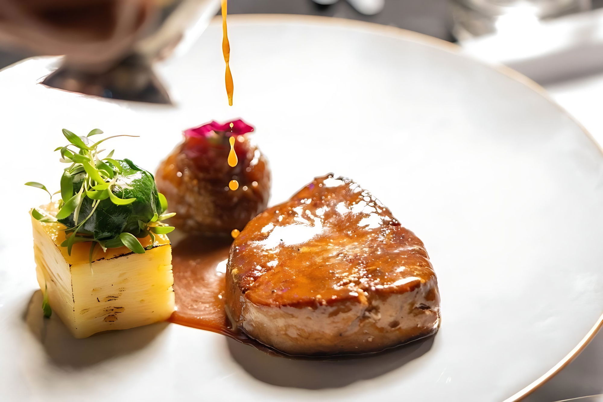 Seared foie gras with a fig, a saffron polenta cube topped with microgreens and a rose petal, sauce being poured over the plate.