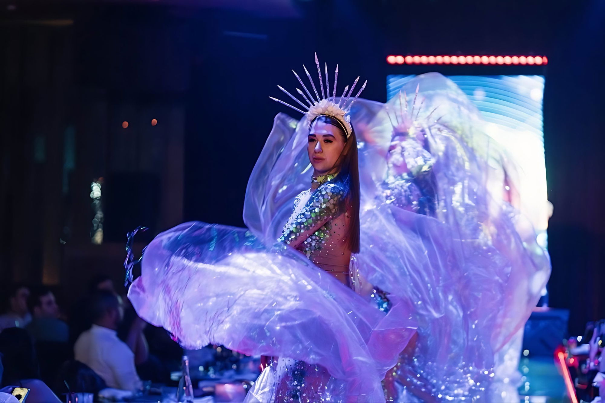 Performer mid-turn in a sequinned costume and spiked headdress, iridescent wings spread, blurred LED screen and seated guests behind.