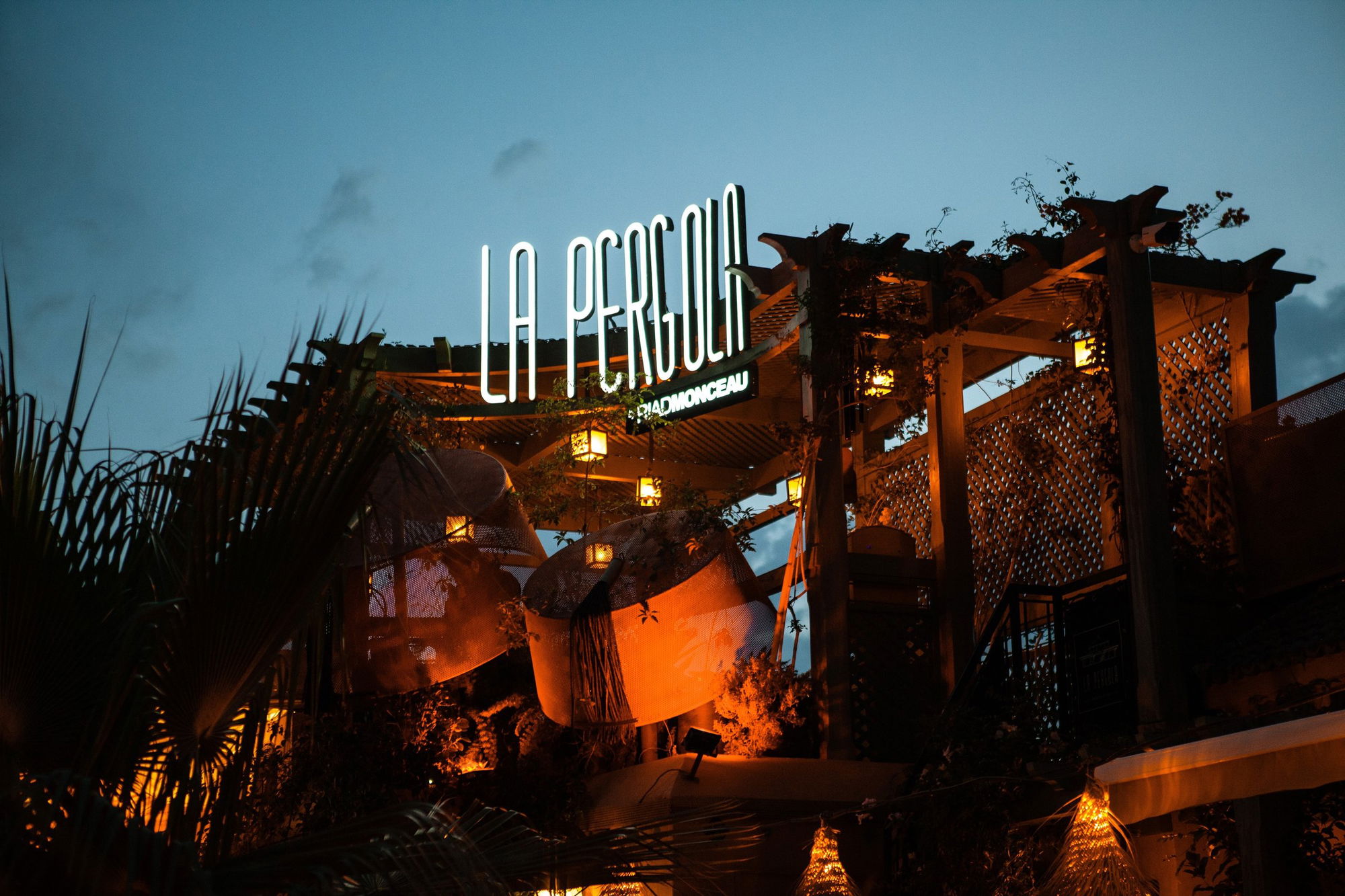 The neon La Pergola sign over the rooftop at dusk, among palms and pavilions