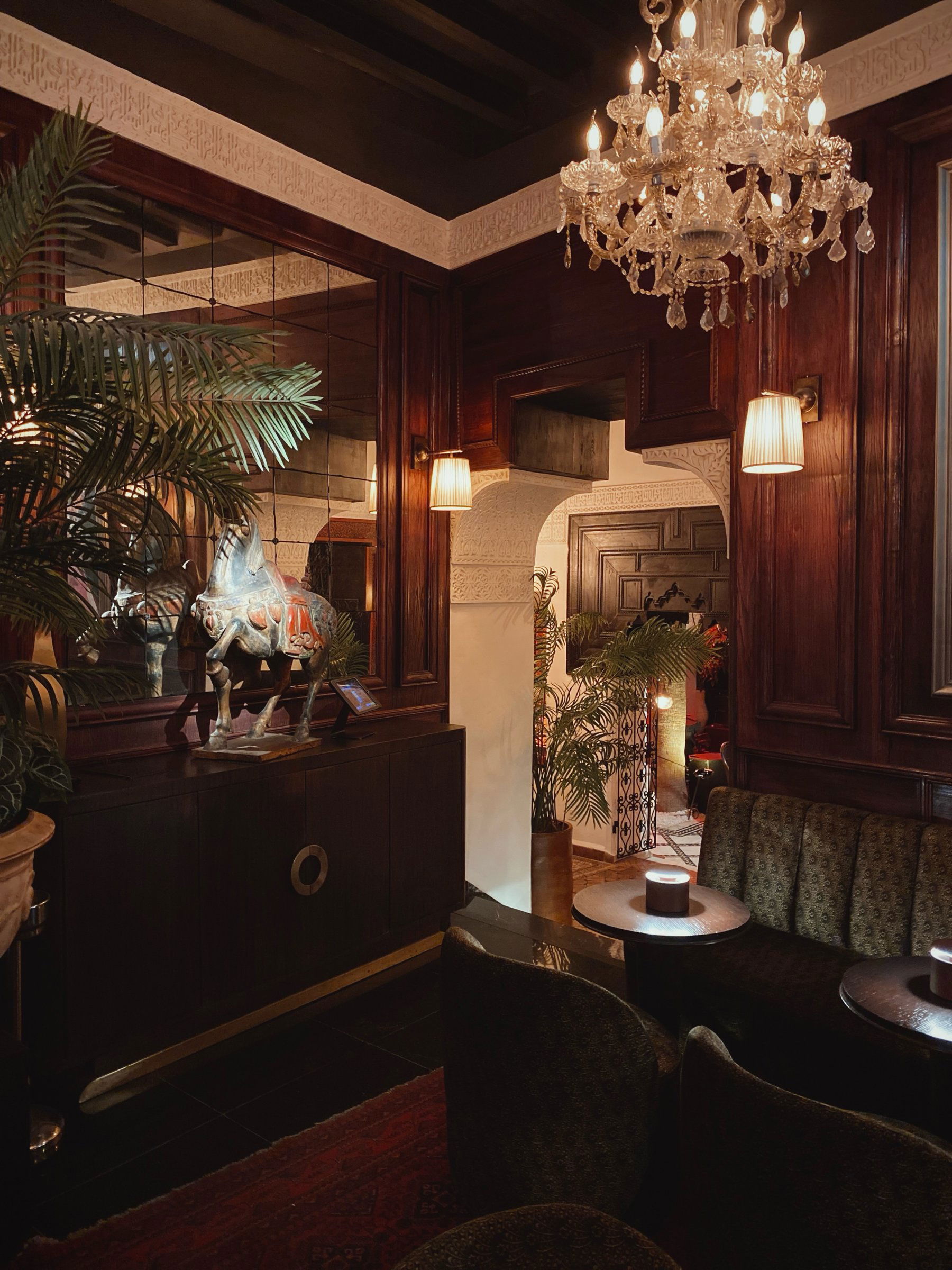 A wood-panelled room with a crystal chandelier, wall lamps and potted palms