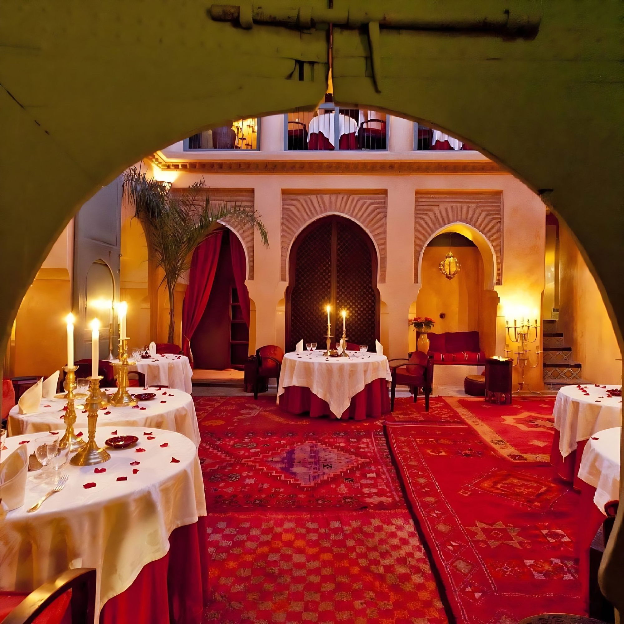 The candlelit courtyard of Le Tobsil seen through an arch, round tables with rose petals on red Moroccan rugs